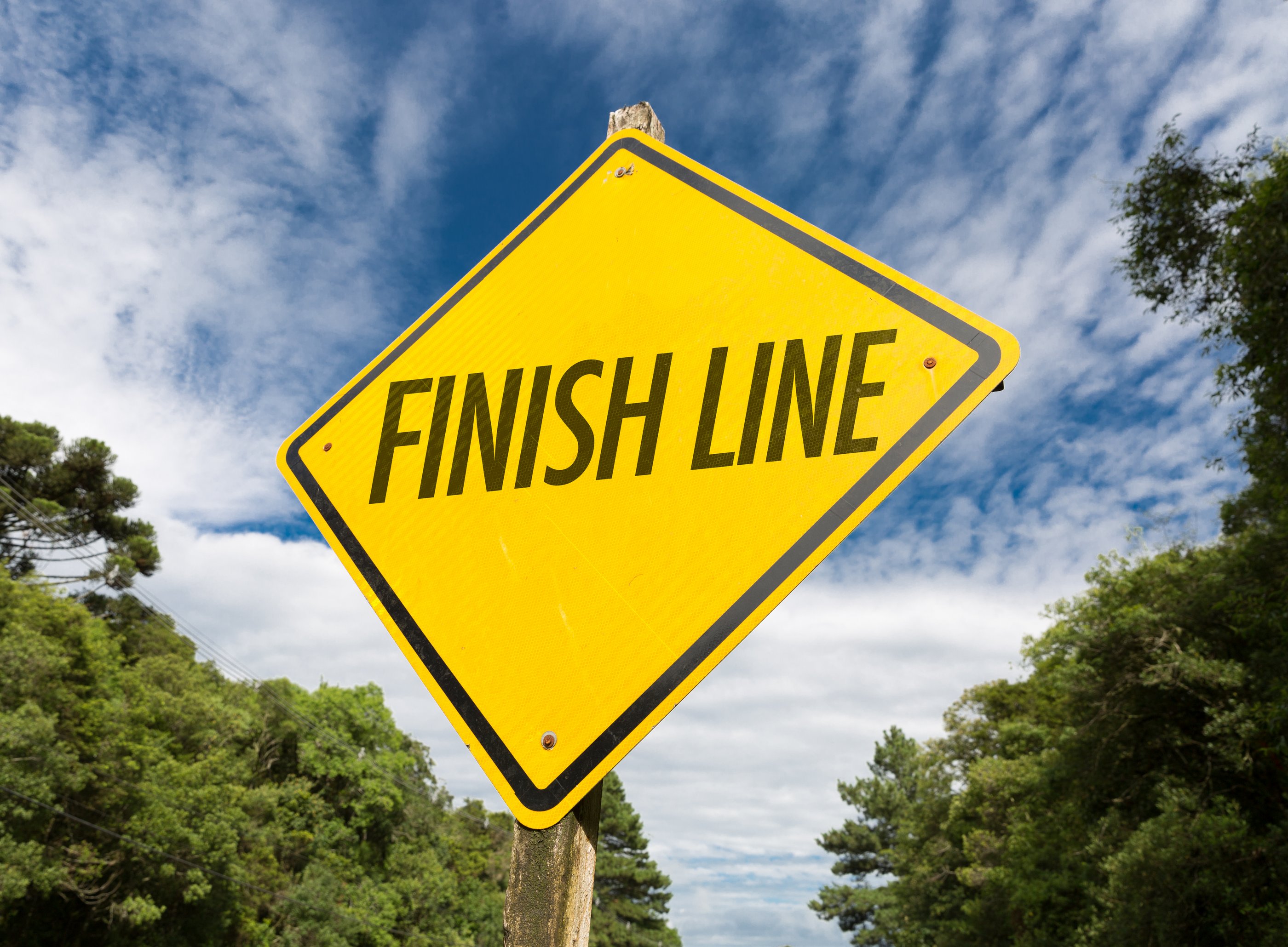 Gelbes Schild mit der Aufschrift Finish Line vor bewölktem Himmel und grünen Bäumen.